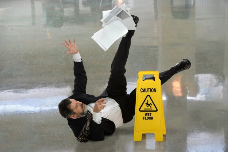 A businessman lands on his back after slipping on a wet floor in front of a caution sign.